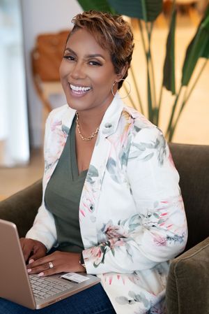 Smiling woman in floral blazer working on laptop while seated, wearing gold jewelry and green top.