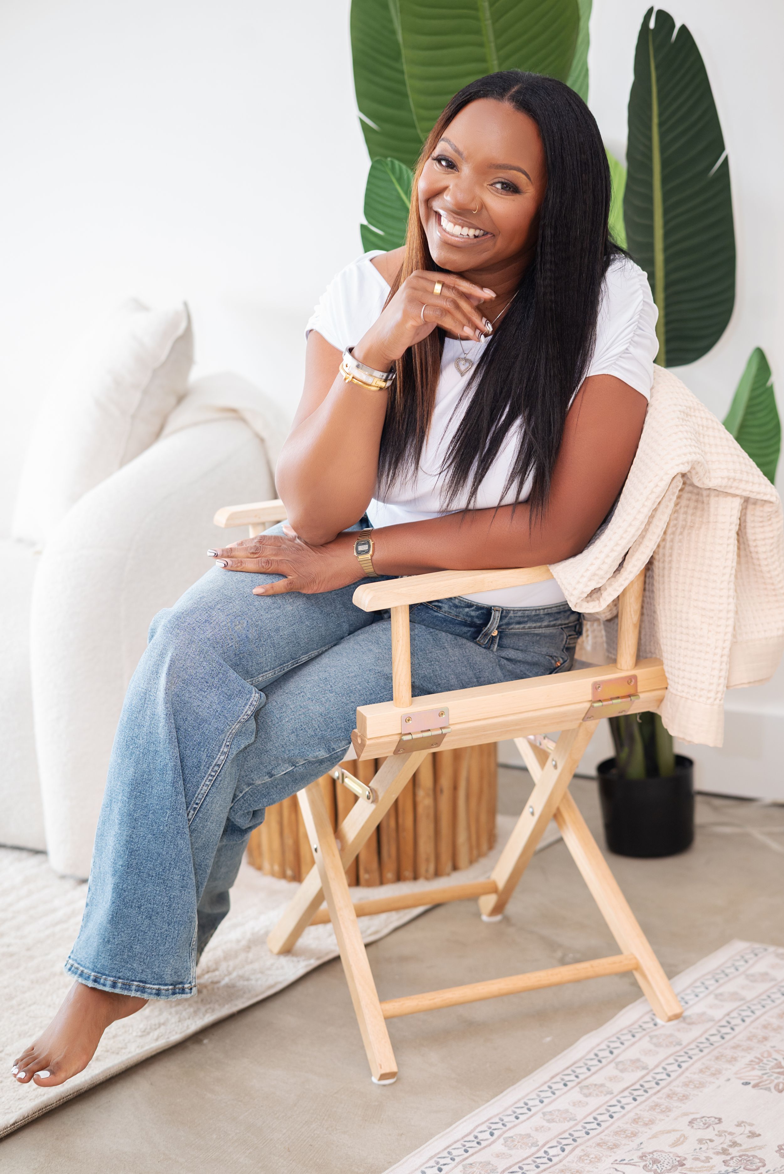 Smiling woman sitting in a wooden folding chair wearing jeans and white tee, posing in a bright modern living space.