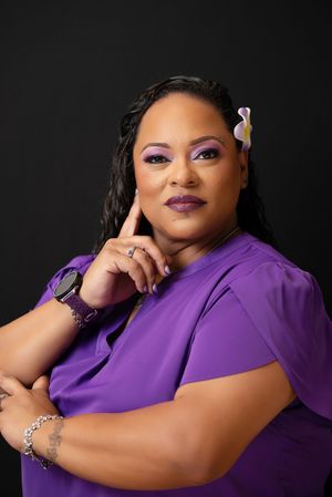 A confident woman in a purple blouse with matching makeup and a flower hair accessory poses against a dark background.