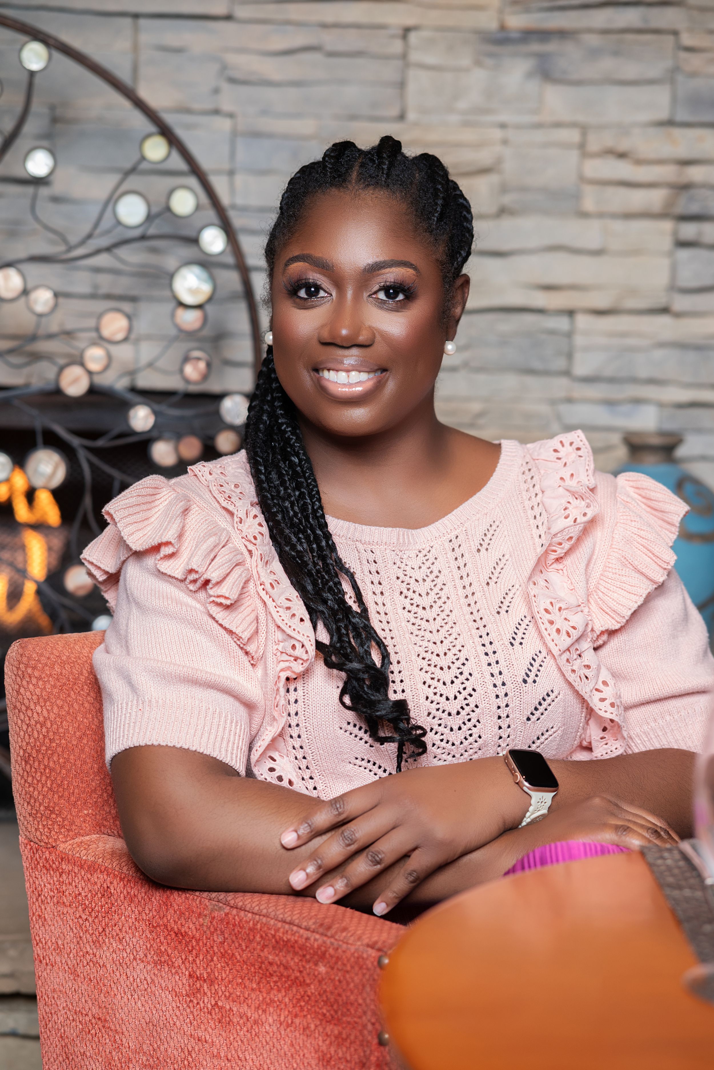 Smiling woman with braids wearing a pink ruffled knit top, seated in an orange chair near a stone fireplace.