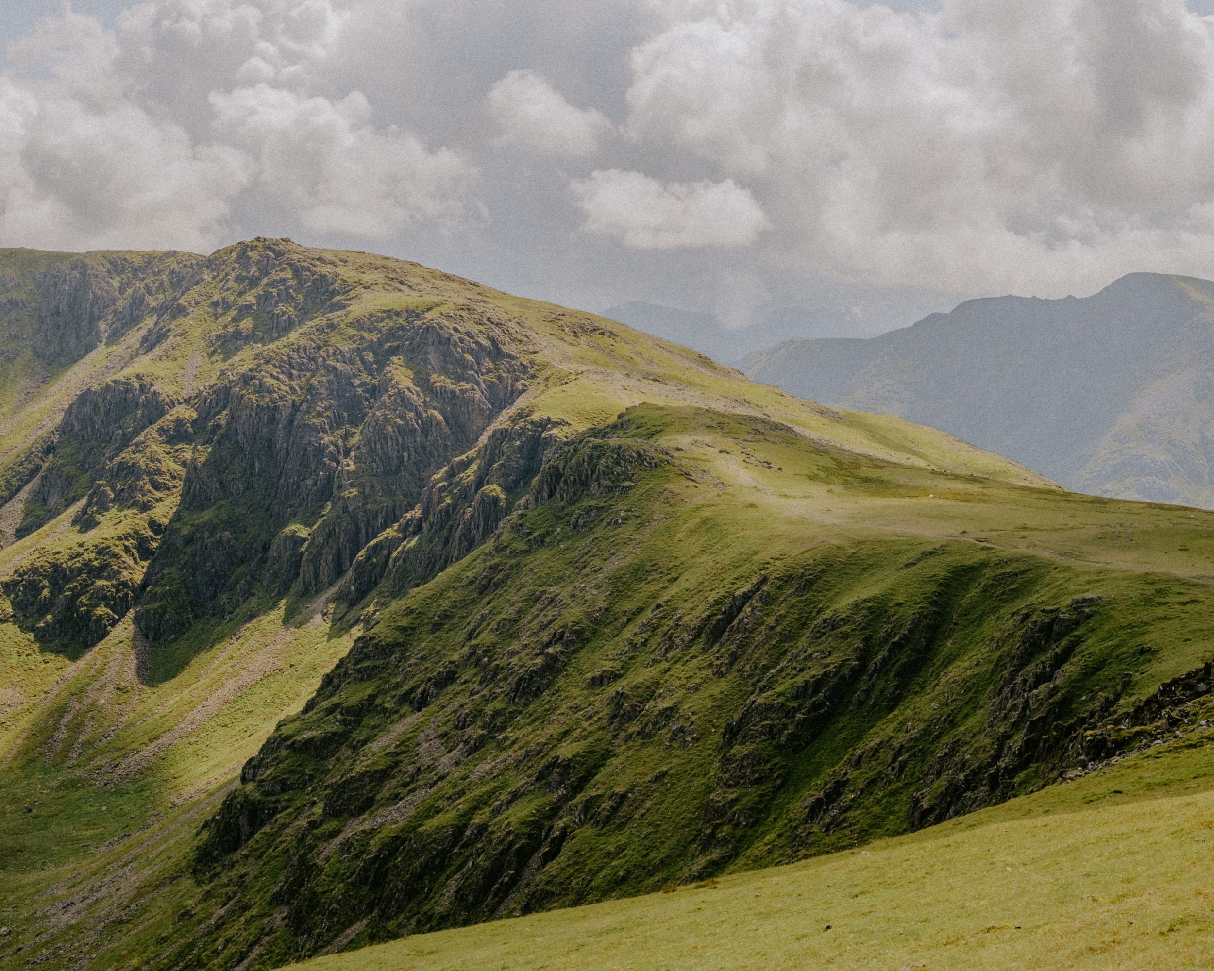 Lush green mountain ridge under cloudy sky with rocky peak and rolling hills in scenic highland landscape.
