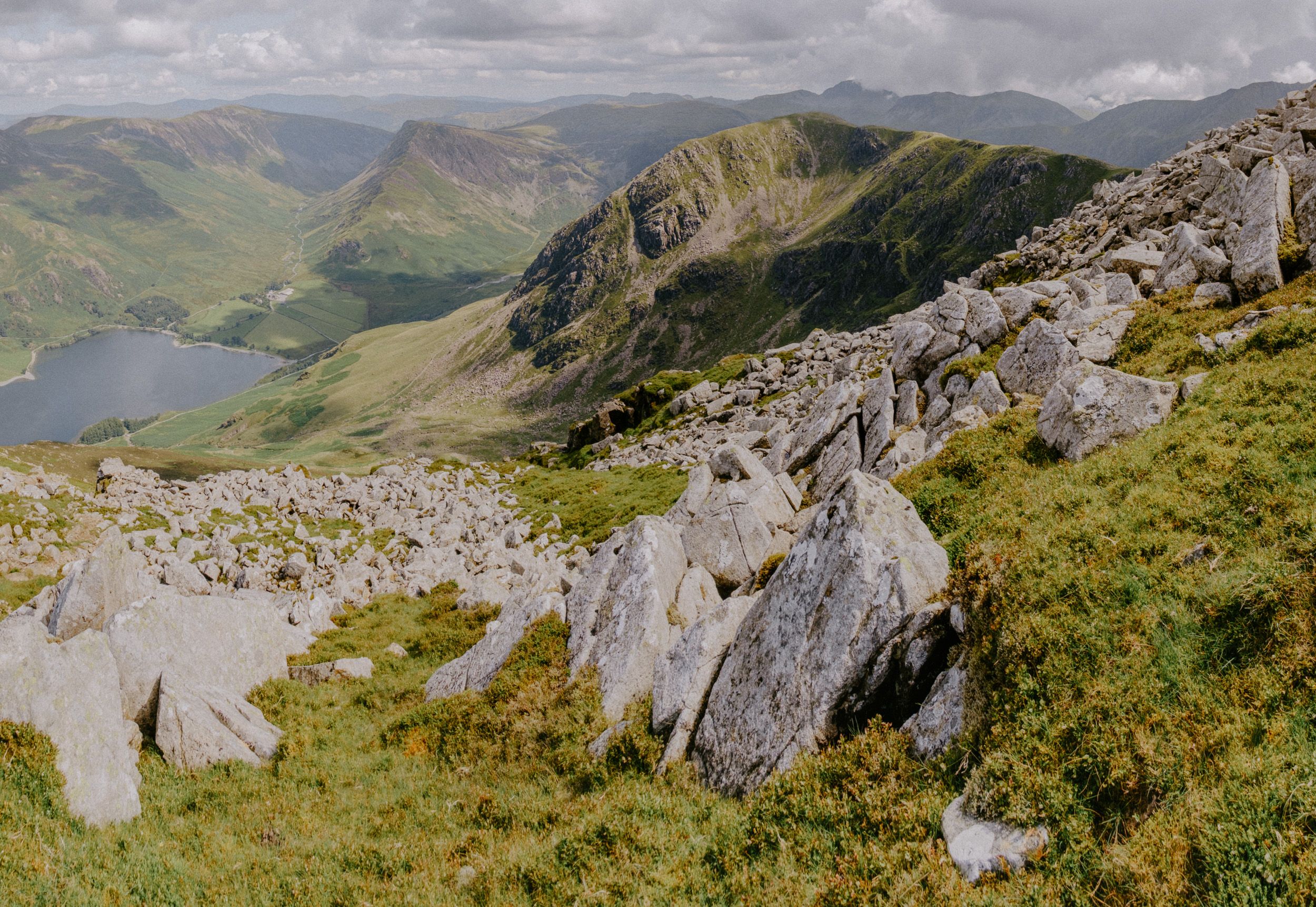 Rocky mountain landscape with a lake in the valley below, surrounded by green hills under a cloudy sky.