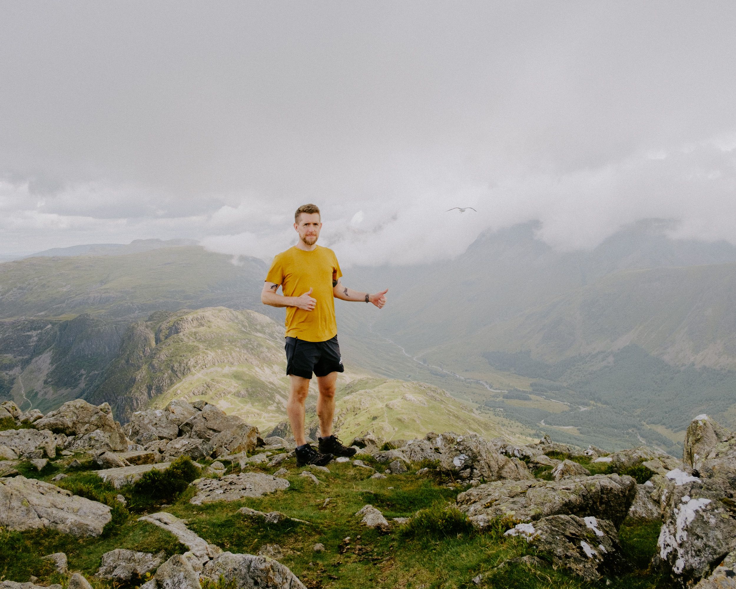Man in yellow shirt standing on a rocky mountain summit with misty green valleys in the background.