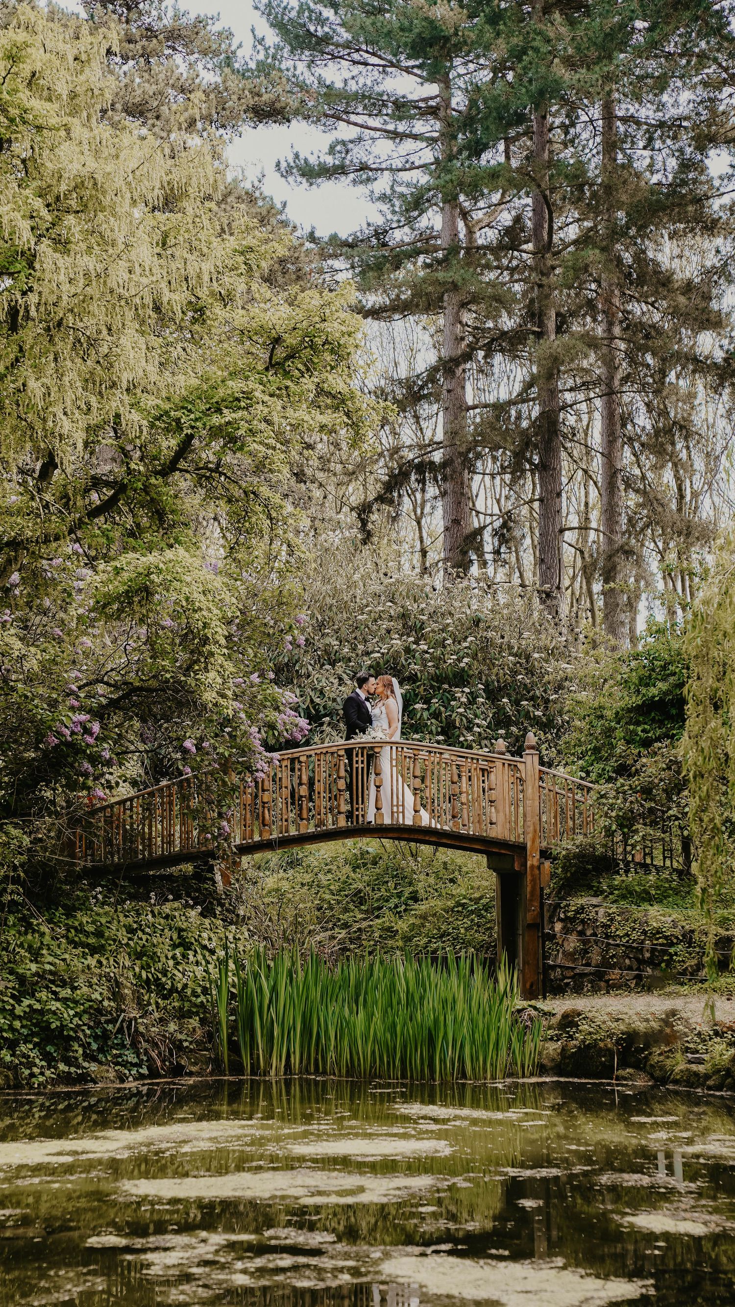 Couple standing on a rustic arched bridge surrounded by lush green trees and a tranquil lily pond in a botanical garden.