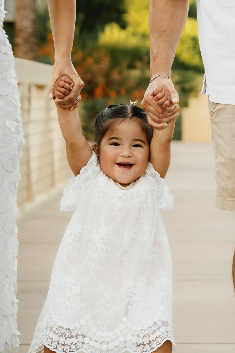 Joyful toddler girl in white lace dress held by parents hands, smiling outdoors in sunny garden setting.