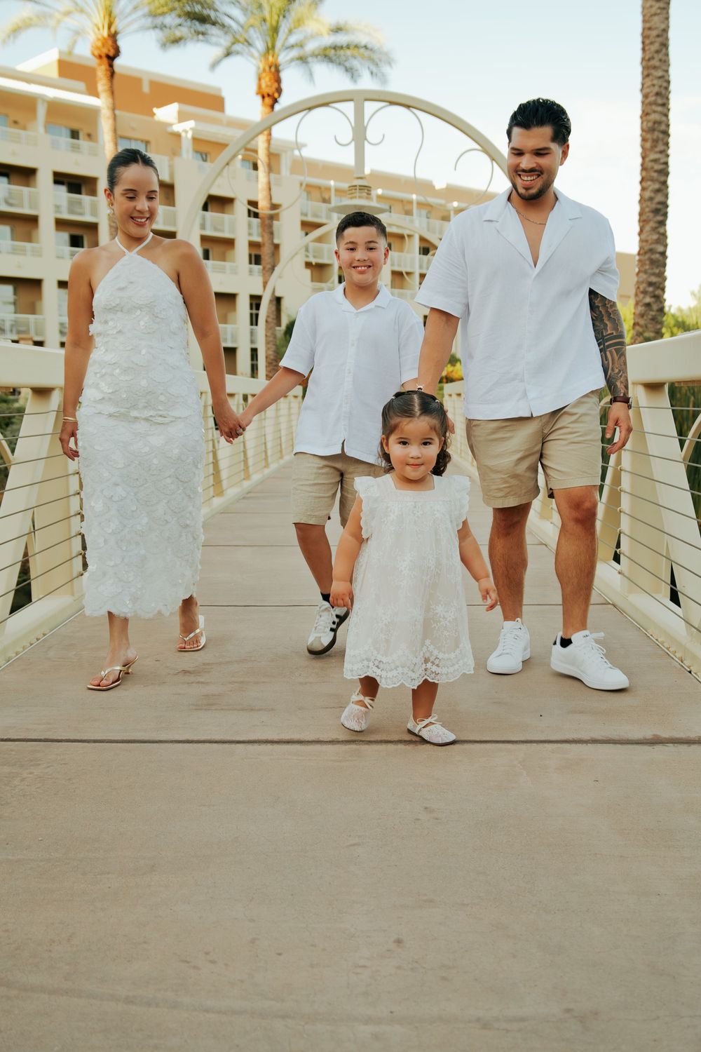 Family of four in white outfits walking on a bridge at a resort, smiling and holding hands.