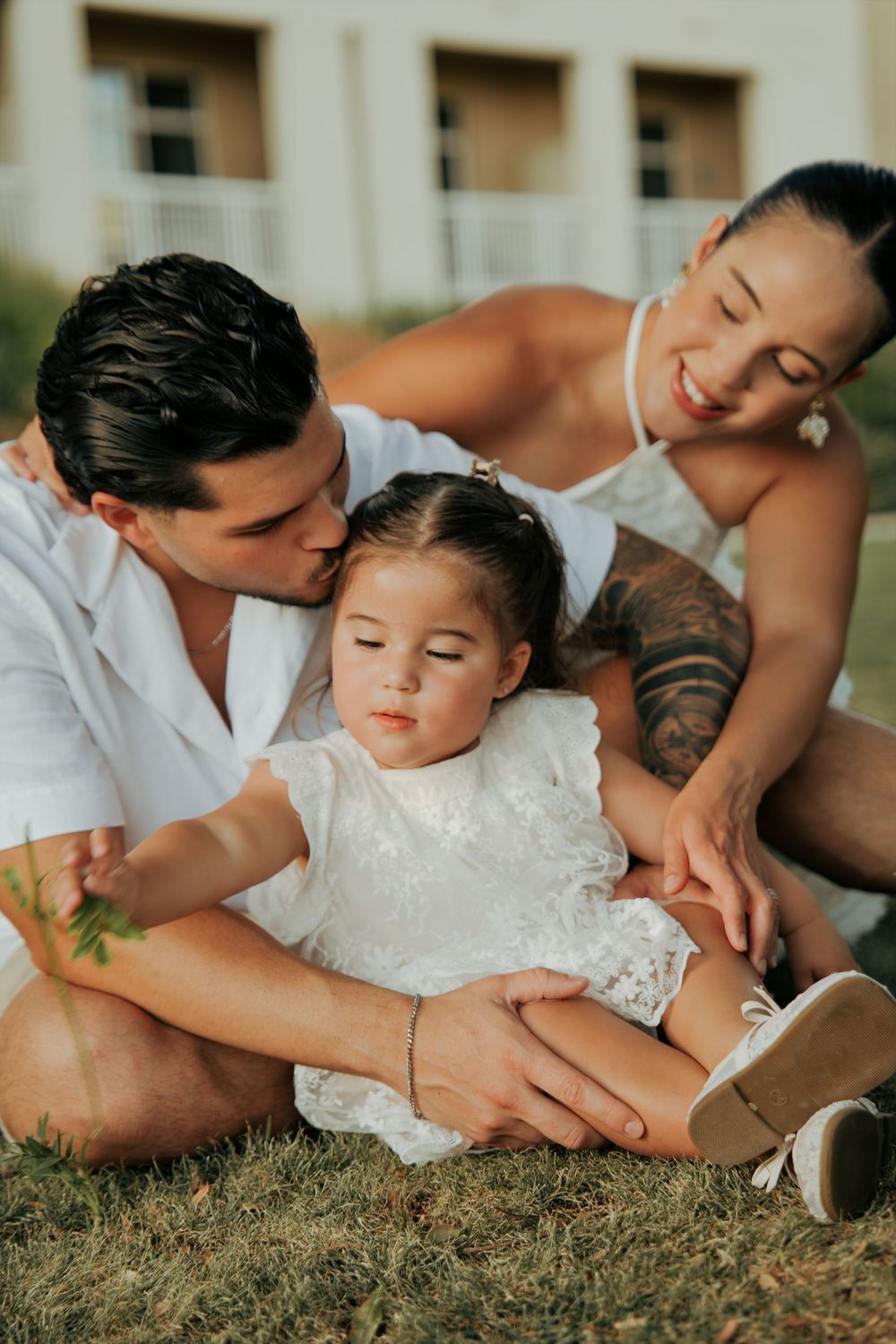 A loving family sits on grass, father kissing toddler girl in white dress, tattooed mother smiling behind them.