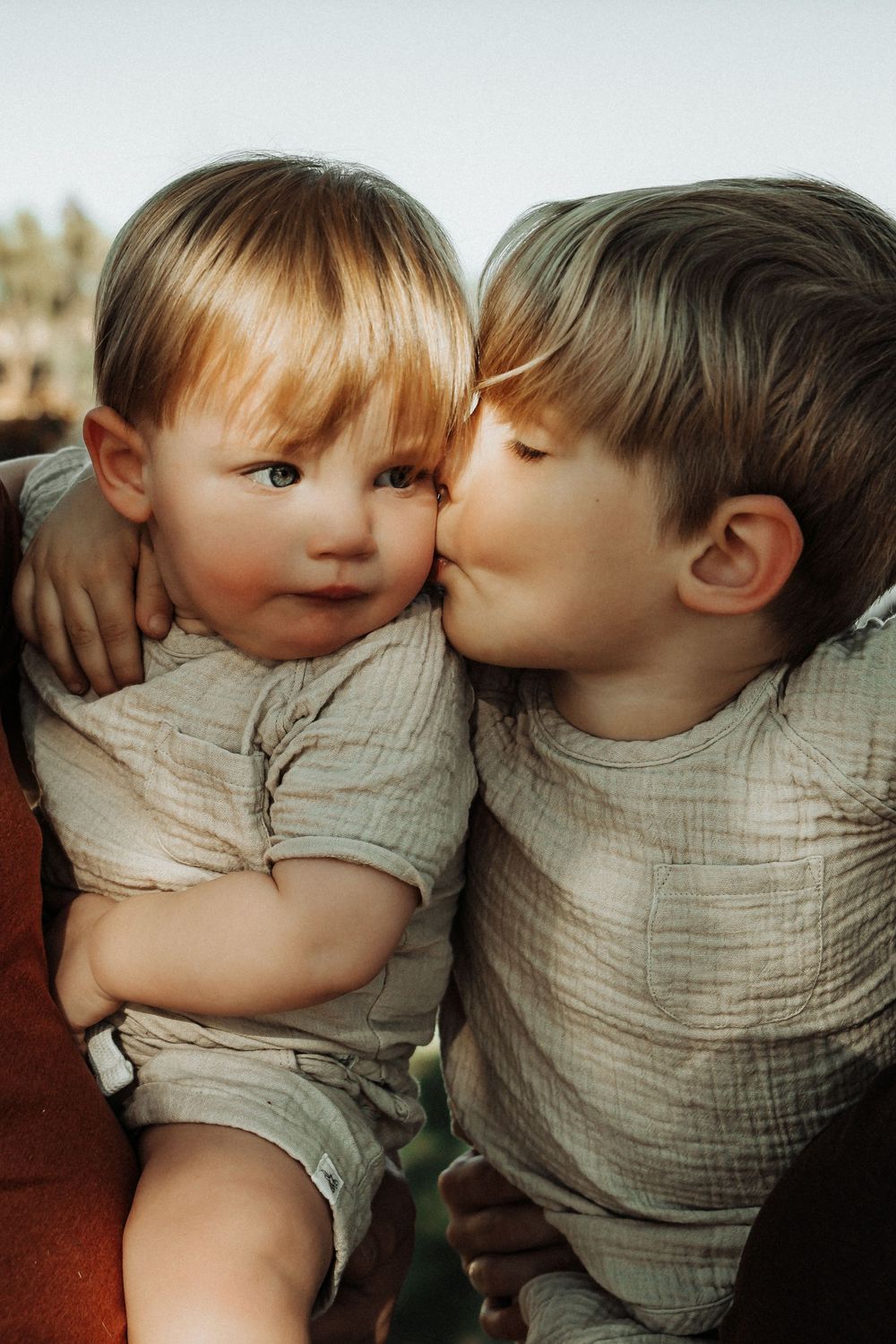 Older boy tenderly kissing younger toddler sibling on the cheek, both wearing matching neutral linen outfits.