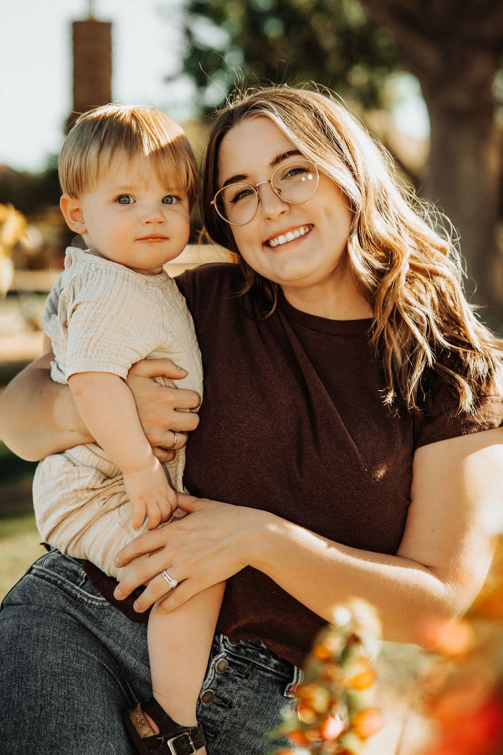A smiling woman with glasses holds a cute toddler outdoors in warm sunlight, both looking at the camera.