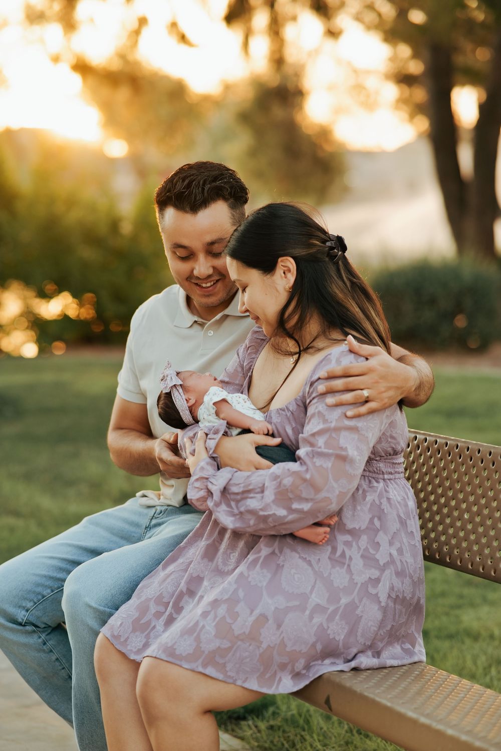 Happy couple sitting on a bench outdoors at sunset, smiling at their newborn baby wrapped in a bow headband.