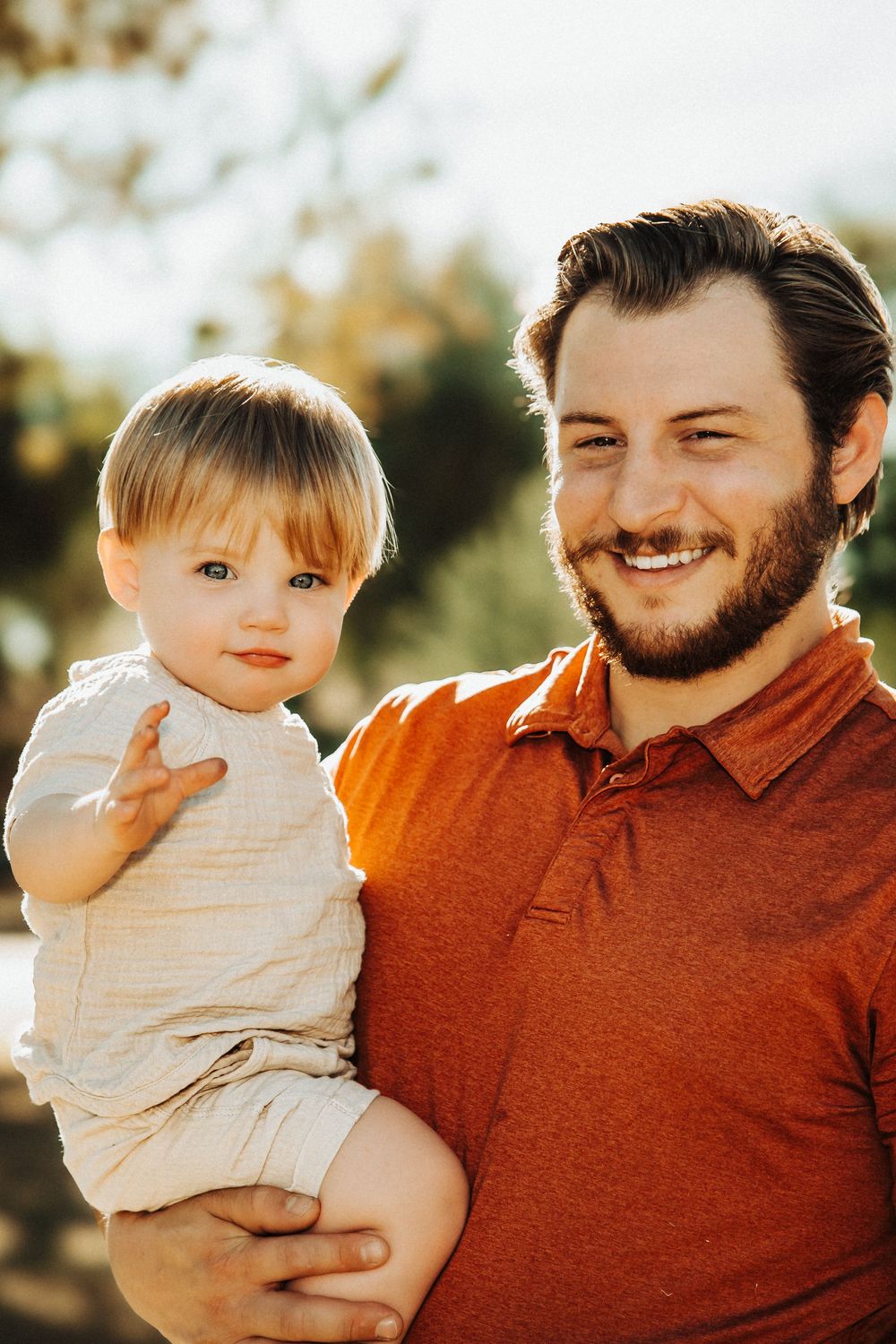 Smiling bearded man in orange shirt holding a happy blonde toddler outdoors in warm sunlight.