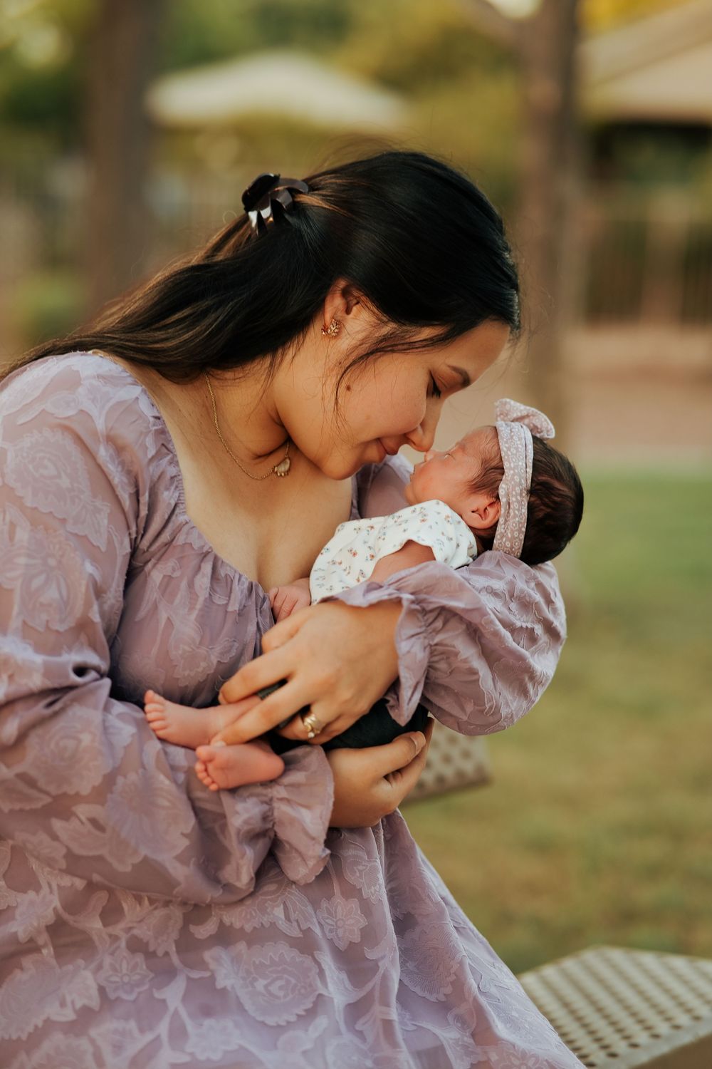 A young mother in a lavender dress tenderly holds her newborn baby outdoors, touching noses affectionately.