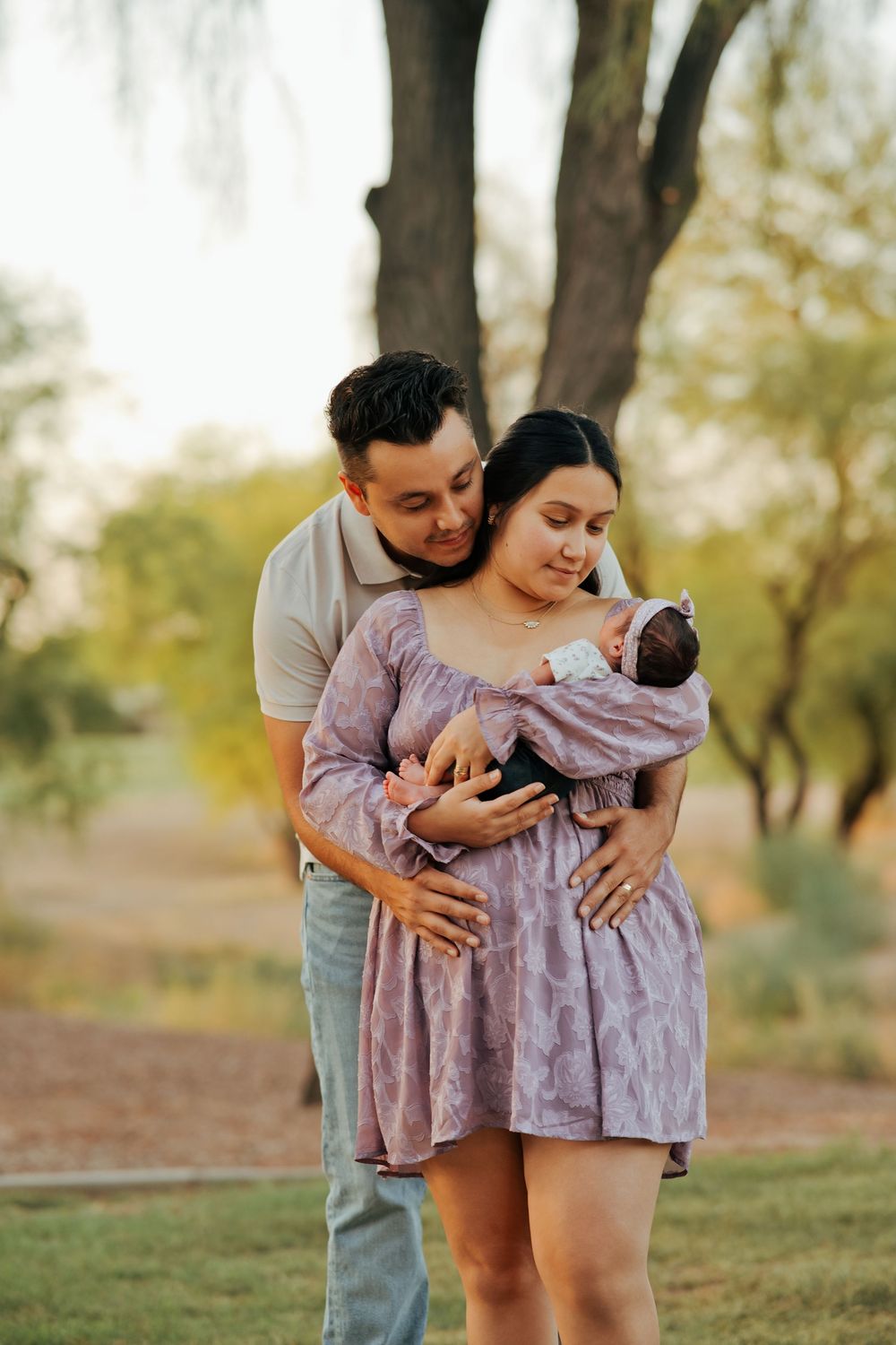 A couple tenderly gazes at their newborn baby outdoors in a park during golden hour.