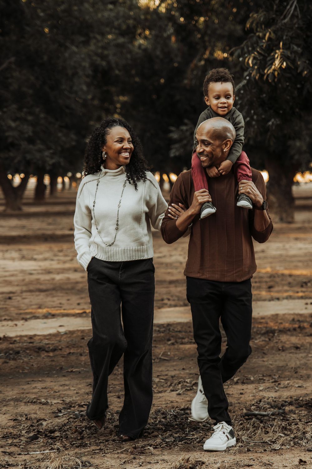 A happy family of three enjoying a walk outdoors, with a man carrying a toddler on his shoulders beside a smiling woman.