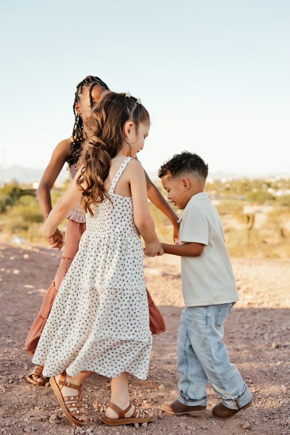 Three children holding hands and playing outdoors in a desert landscape during golden hour.
