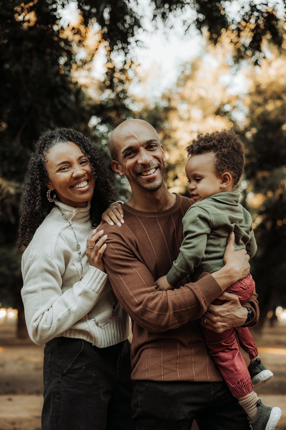 Happy Black family with parents smiling and holding toddler in autumn park setting.