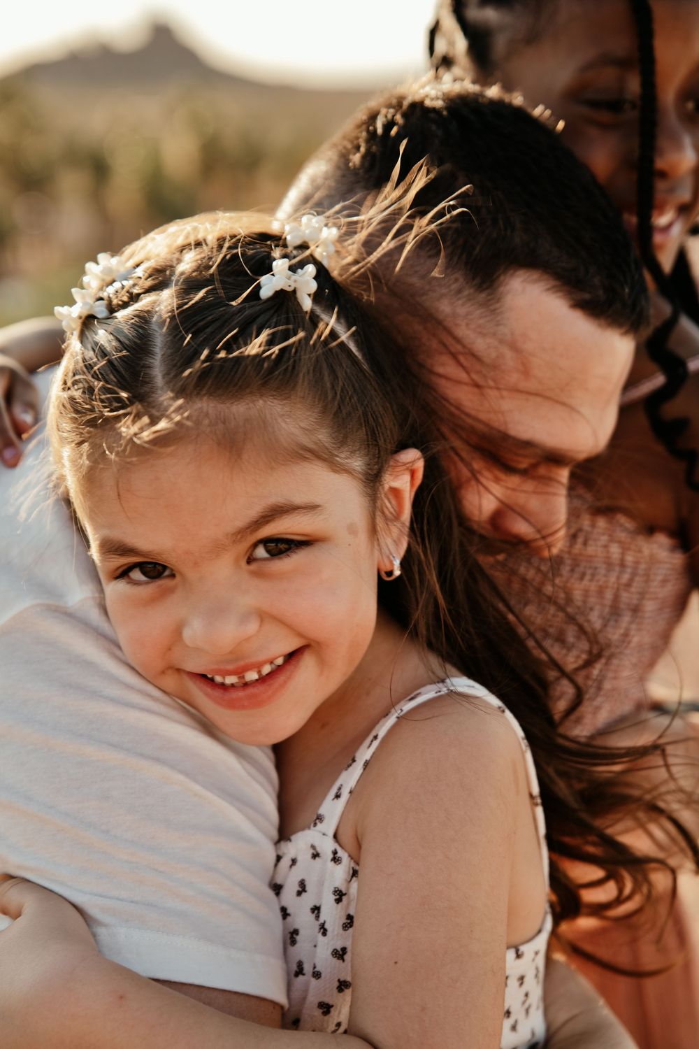 A smiling young girl with flower hair accessories hugs someone outdoors in warm golden sunlight.