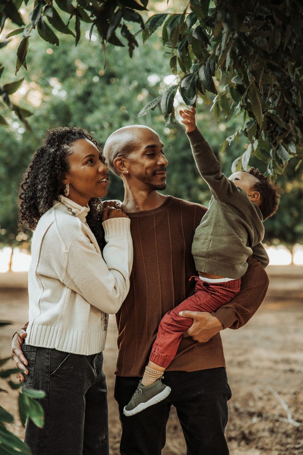 A happy Black family outdoors, father holding toddler reaching for leaves, mother smiling beside them in a green park.