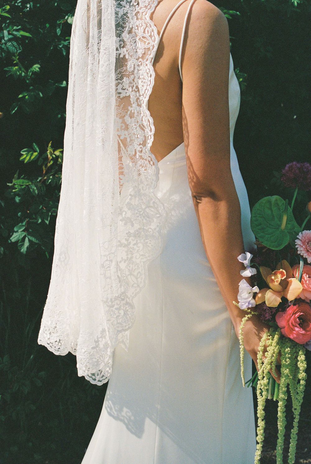 Bride in elegant backless lace wedding gown with cathedral veil holding a vibrant colorful bouquet outdoors.