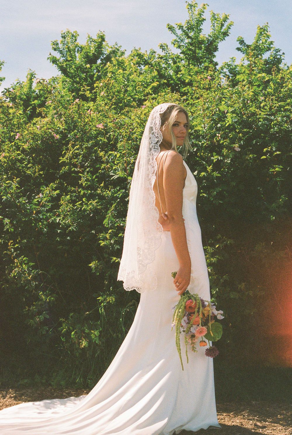Bride in white lace wedding gown and veil holding floral bouquet, posing outdoors with green trees background.