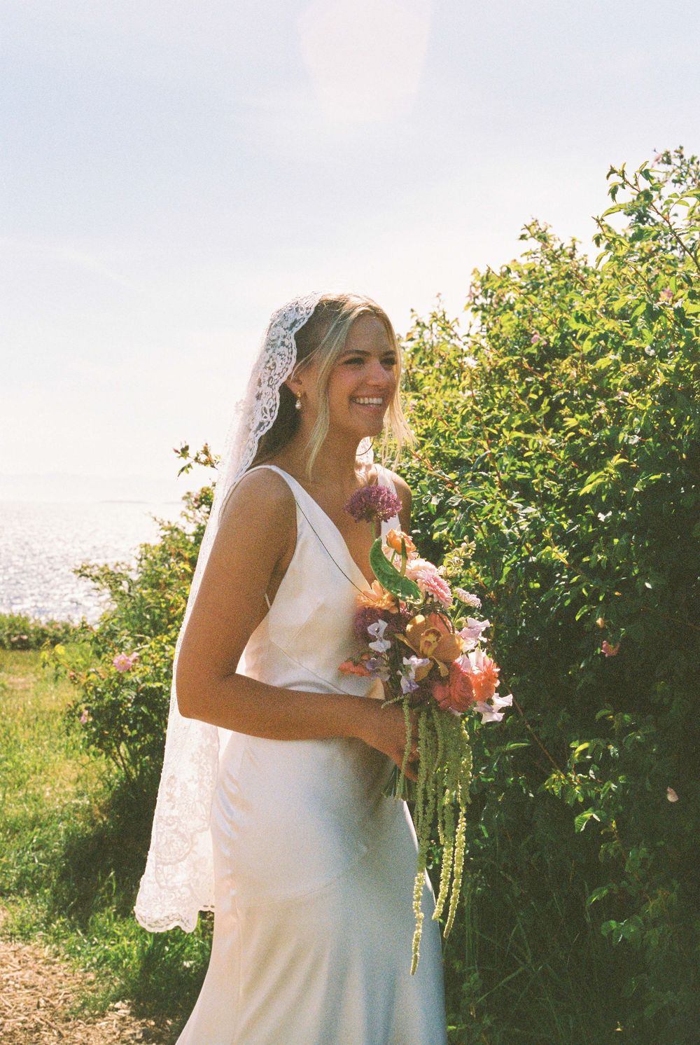 Smiling bride in white gown and lace veil holds colorful bouquet outdoors with ocean view and green hedges.