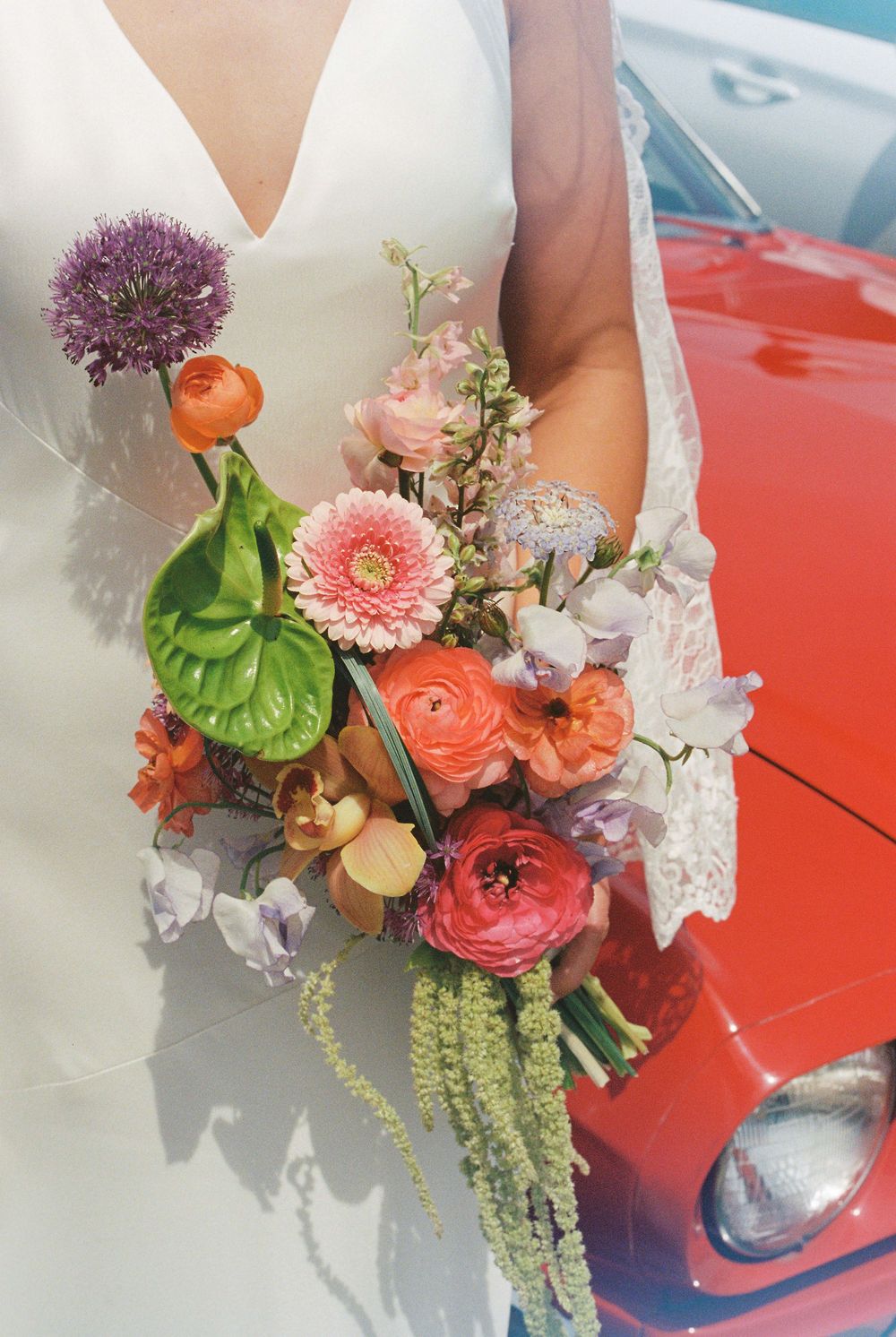 Bride in white lace gown holding vibrant wedding bouquet with ranunculus, gerbera daisies, and allium near red car.