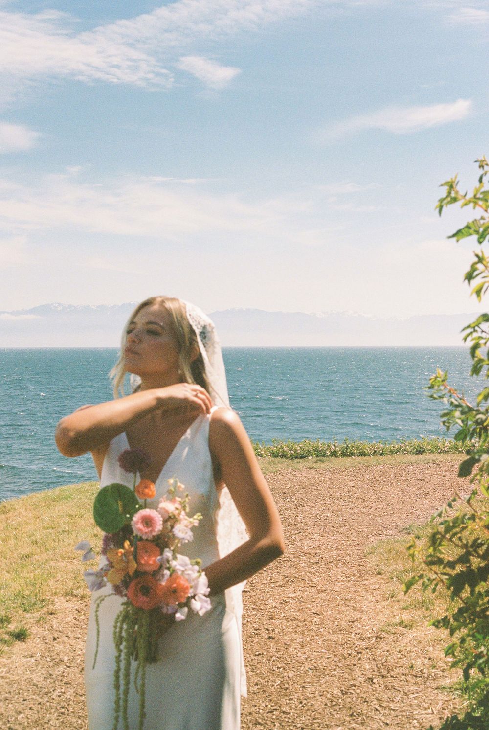 Bride in white gown and veil holds colorful bouquet by ocean cliffside under blue sky.