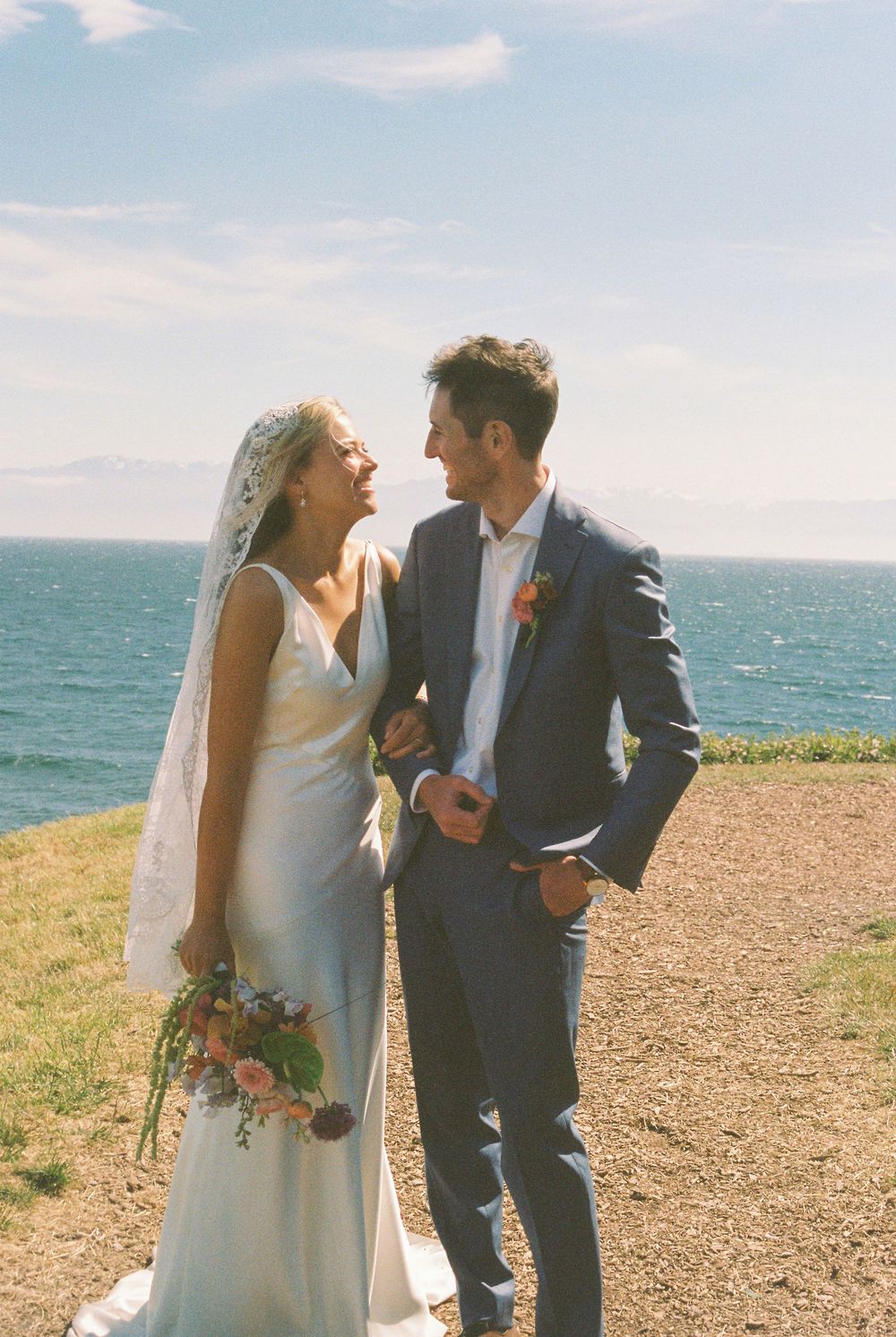 Bride and groom laugh together on coastal cliffside, she holds bouquet, he wears navy suit.