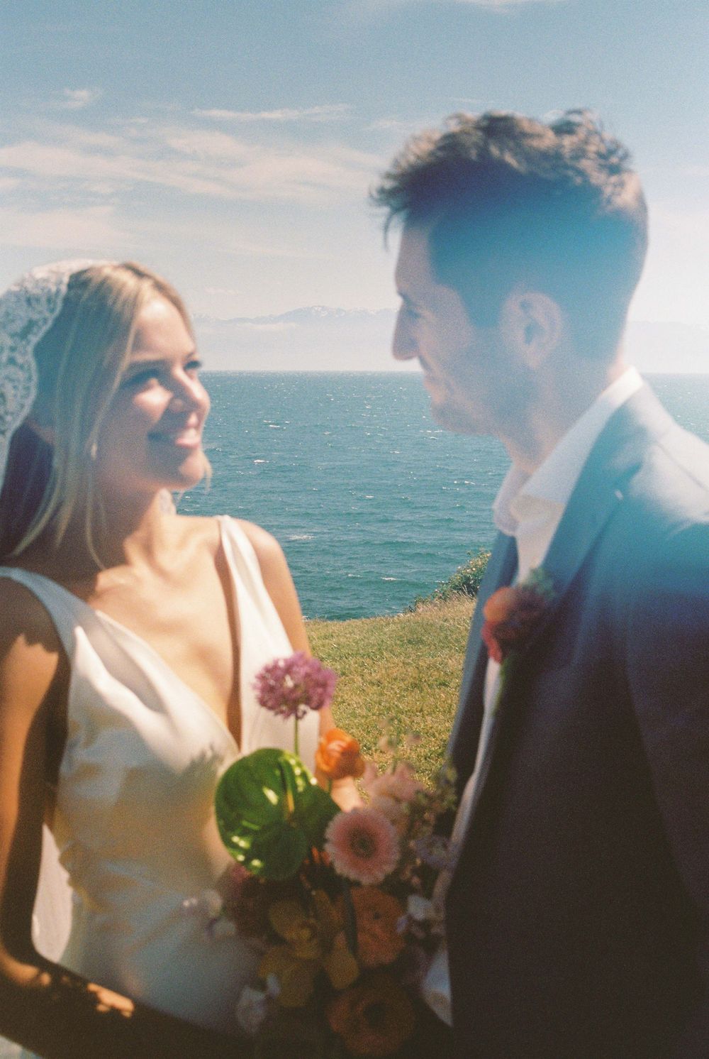 Bride and groom share a romantic moment by the ocean, with colorful bouquet and scenic coastal backdrop.