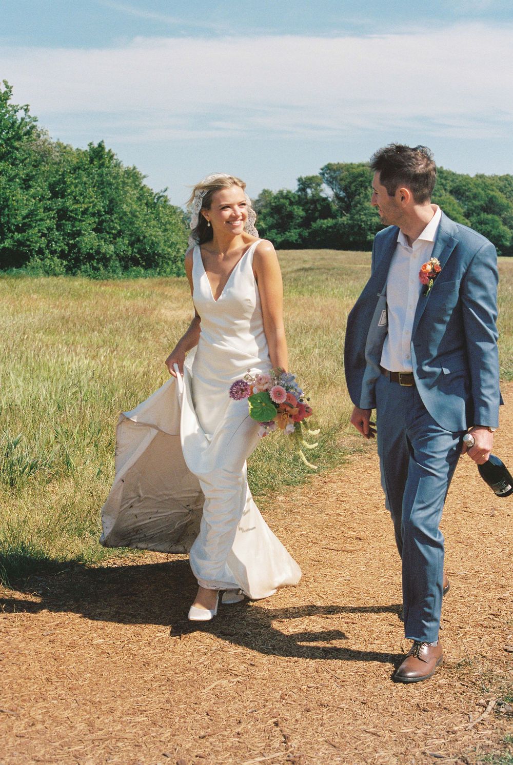 Bride and groom walking on a sunny dirt path, she holds a colorful bouquet in a white gown, he wears a blue suit.