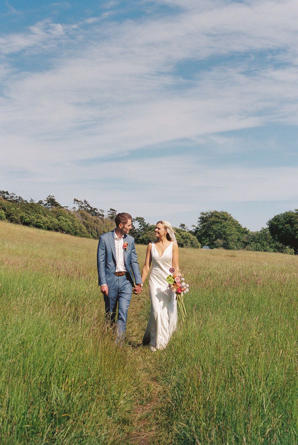 Bride and groom holding hands walking through a lush green meadow under a blue sky on their wedding day.
