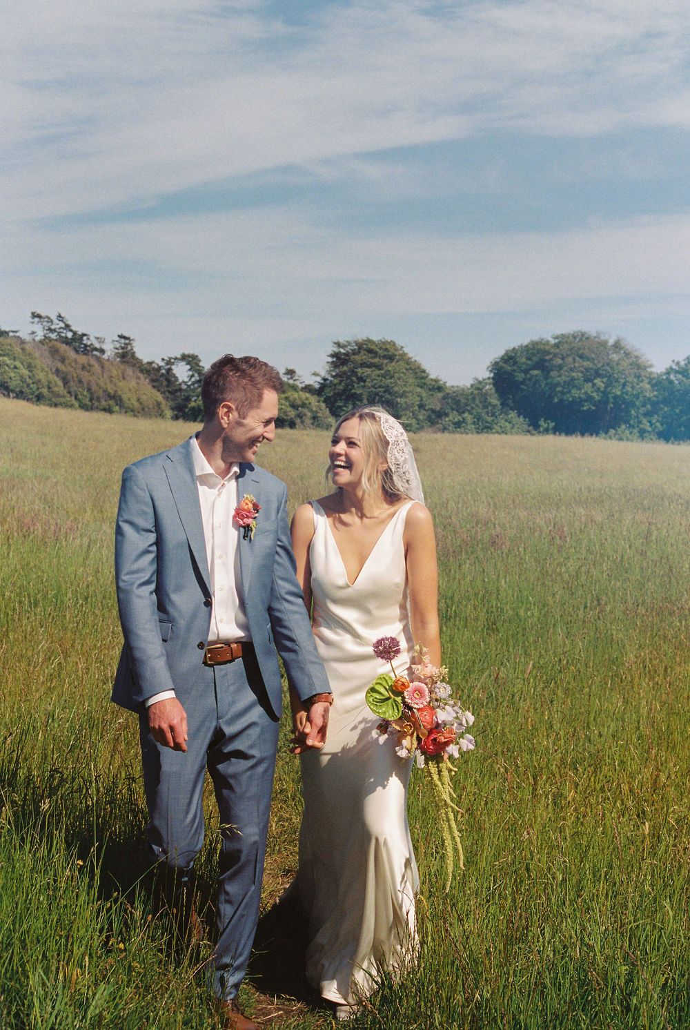 Laughing bride in white gown and groom in blue suit hold hands in a sunlit green field on their wedding day.