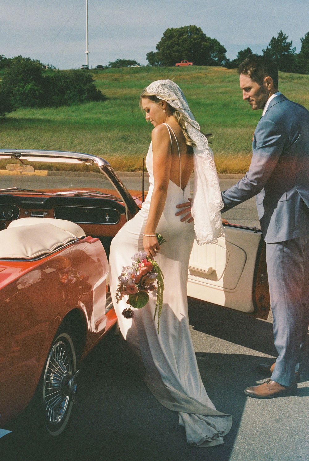 Bride in white gown and veil steps out of a classic red convertible assisted by groom in blue suit.