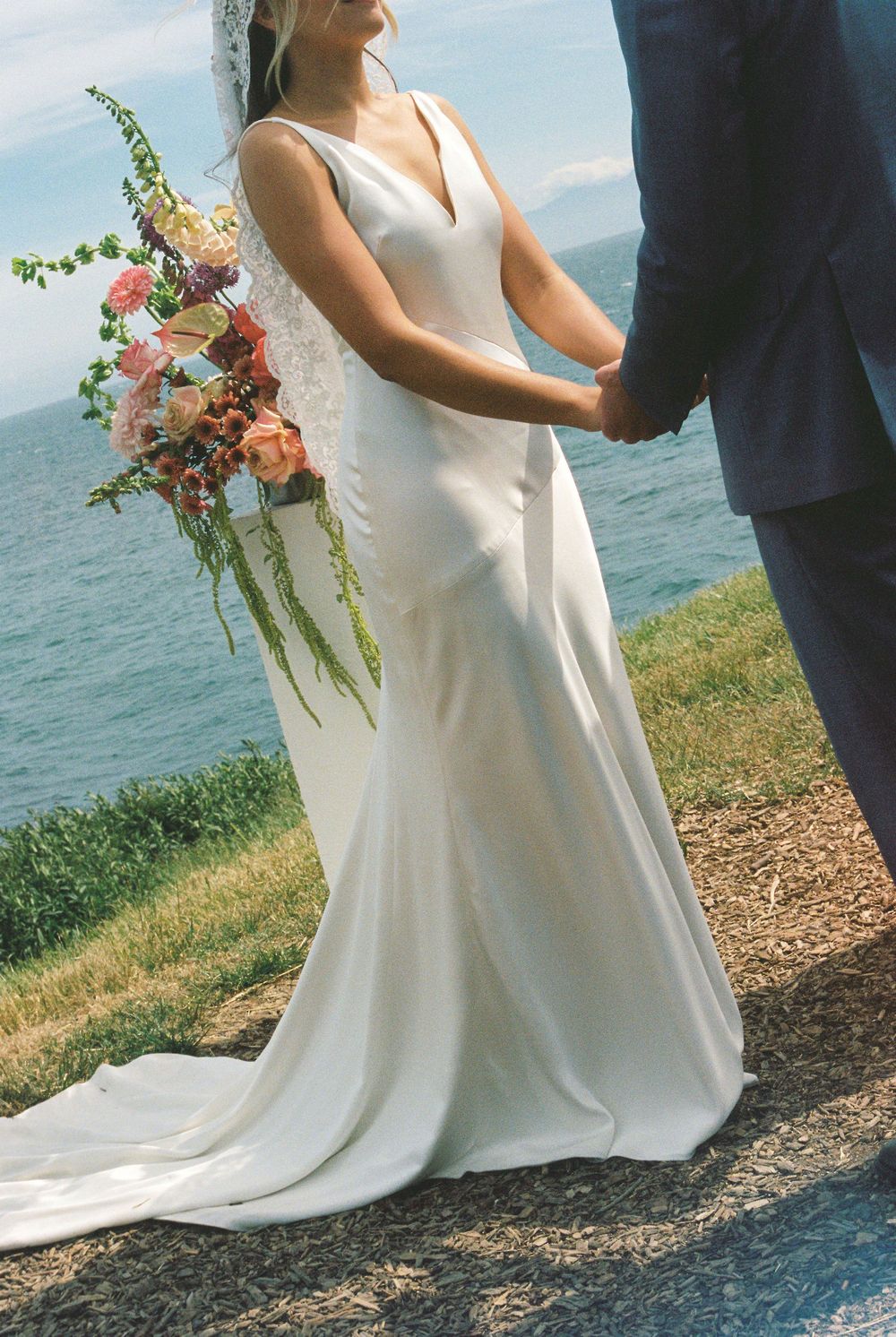Bride in elegant white satin wedding gown holding colorful bouquet, standing at outdoor coastal ceremony with ocean views.