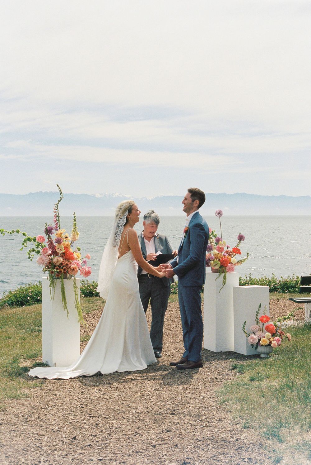 Couple exchanges vows outdoors by a lake with colorful floral arrangements on white pedestals, officiant behind them.