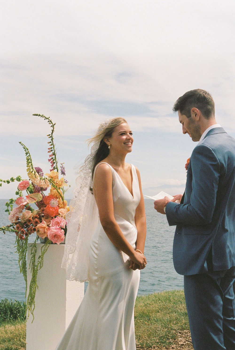 Bride in white gown smiling at groom reading vows outdoors by the water with colorful floral arrangement.