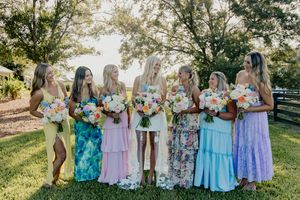 Bride with bridesmaids in colorful mismatched dresses holding bouquets outdoors on a sunny day.