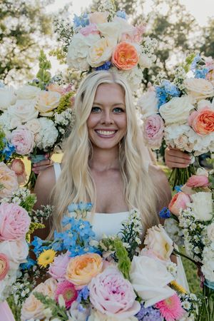 Smiling blonde bride framed by a large colorful floral arrangement with roses and blue flowers.