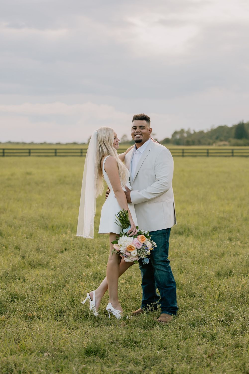 Bride and groom embracing in a green field with a wooden fence in the background on their wedding day.