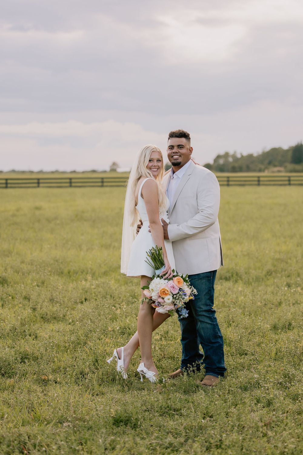 Smiling couple poses in green field at golden hour, bride holding colorful wildflower bouquet.