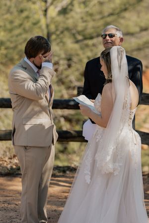Emotional groom wiping tears during outdoor wedding ceremony while bride reads vows, officiant smiling behind them.