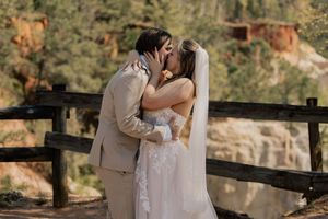 Bride and groom share first kiss at outdoor wedding venue with rustic wooden fence and forest backdrop.