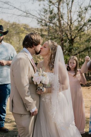 Bride and groom kiss surrounded by bubbles while wedding guests celebrate in sunny outdoor ceremony.