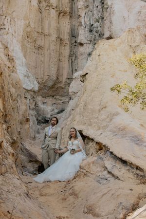 Bride and groom pose together in dramatic sandy canyon walls wearing elegant wedding attire.