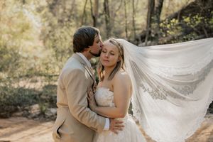 Groom tenderly kisses bride on cheek while her lace veil billows in forest wedding portrait.