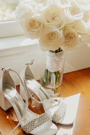White pearl bridal heels beside a bouquet of white roses in a glass vase.