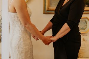 Two women in wedding attire hold hands tenderly during an intimate ceremony.