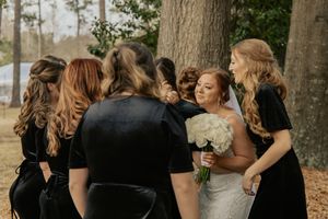 Bride holding white bouquet surrounded by bridesmaids in black dresses hugging outdoors near trees.
