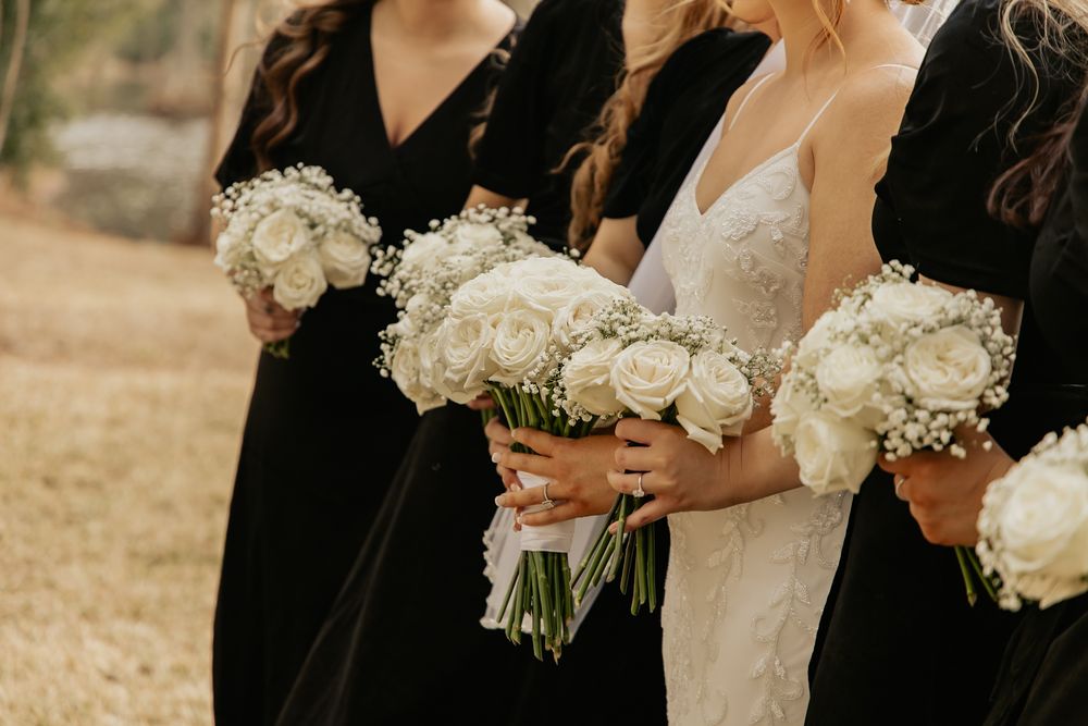 Bride and bridesmaids in black dresses holding white rose and baby breath bouquets.