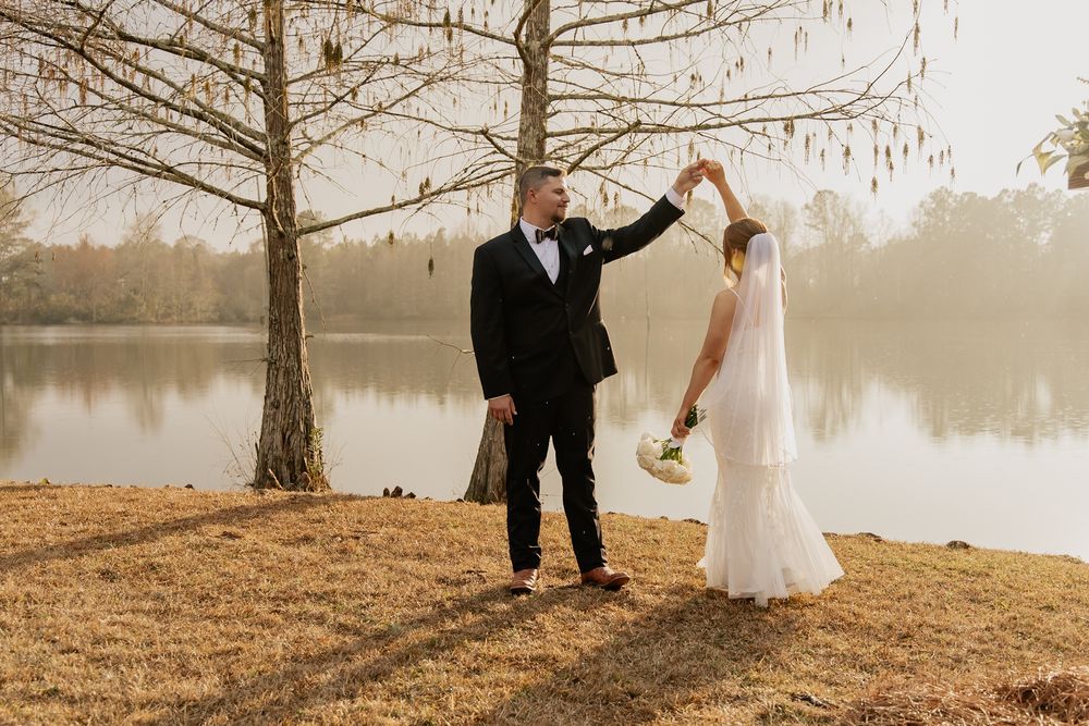 Groom twirling bride in white gown near misty lake with bare trees in autumn setting.